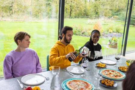 Un joyeux rassemblement d'amis autour d'une table à manger, dégustant une gamme de plats appétissants et du vin dans un cadre de serre rempli de lumière. La toile de fond verte luxuriante vue à travers les murs de verre ajoute un