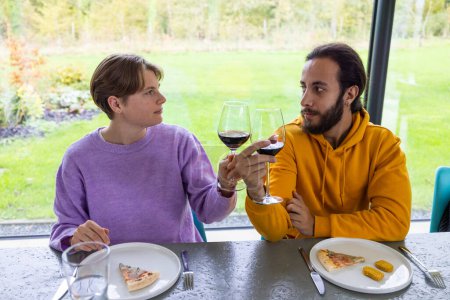 Dieses einladende Foto fängt zwei Freunde ein, die sich einen Moment beim Wein austauschen und bedeutungsvolle Blicke austauschen, die auf ein tiefes Gespräch und Verständnis hindeuten. Das ungezwungene Dining-Erlebnis mit Pizza-Scheiben
