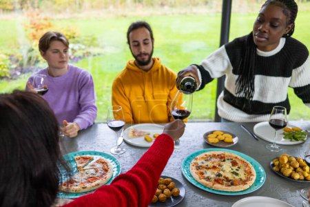 Una instantánea de la diversidad moderna, esta imagen representa a un grupo de amigos multiculturales de varios géneros y edades disfrutando de una comida agradable. Están reunidos en un comedor con vistas al jardín, compartiendo