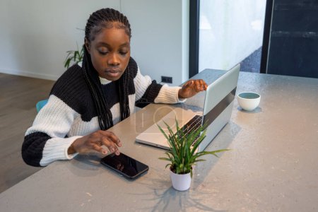 Cette image montre une jeune femme afro-américaine en pull noir et blanc, parfaitement multitâche dans un espace de travail minimaliste. Elle exploite un smartphone et un ordinateur portable simultanément, suggérant