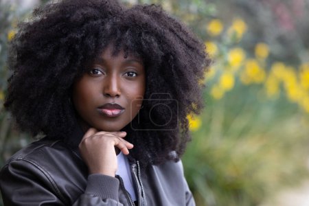 A thoughtful African American woman with voluminous afro hair and a black leather jacket poses against a blurred natural background with yellow flowers, conveying introspection and style