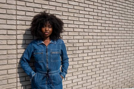 An African American woman with natural afro-textured hair stands confidently against a white brick wall backdrop. She sports a stylish full denim jumpsuit that cinches at the waist, complementing her