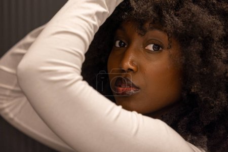 Dieses Bild zeigt eine Nahaufnahme einer afroamerikanischen Frau mit auffallenden Afrohaaren, die ein lässiges weißes Oberteil trägt. Ihre Arme sind anmutig positioniert und umrahmen ihr nachdenkliches Gesicht, das gegen ein weiches