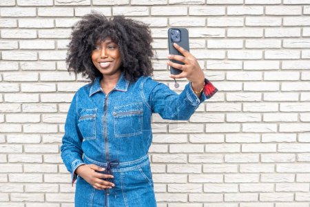 Captured against a textured white brick wall, a vibrant African American woman with a lush afro smiles as she takes a selfie. She sports a trendy denim jumpsuit, red-cuffed sleeves adding a pop of