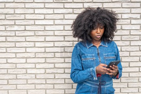 A young African American woman stands focused on her smartphone against a white brick wall backdrop. Her curly afro frames her face while her denim outfit reflects a casual, urban style, depicting a