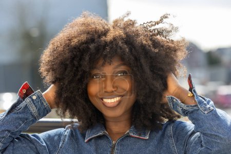 Ein lebendiges Porträt einer lächelnden Afroamerikanerin mit einem üppigen Afro, der die Natur genießt. Das Jeansgewand ergänzt ihr fröhliches Auftreten unter freiem Himmel und steht beispielhaft für eine freigeistige
