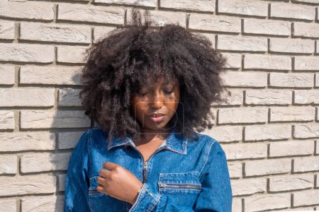 Captured in contemplation, a young African American woman with a full, natural afro stands against a stark white brick wall. The sunlight highlights her denim jacket, adding warmth to the image, while