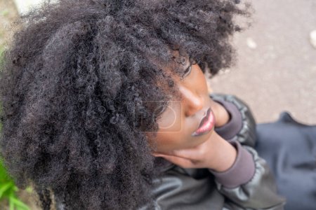 An intimate perspective of a young African American woman looking upwards, her voluminous afro framing her dreamy expression. A sense of wonder and daydreaming is captured against a blurred urban