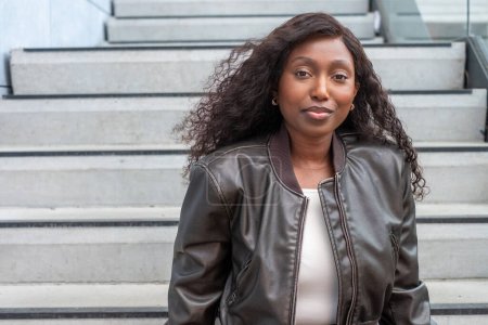 An elegant Black woman sits comfortably on concrete stairs, her posture relaxed and confident. She wears a chic leather jacket over a white top, her hair flowing naturally. The urban setting and her