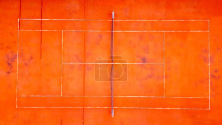 An aerial perspective showcases the geometric beauty of a vibrant orange tennis court, the bold white lines creating a stark contrast. The surface shows signs of weathering, adding texture and