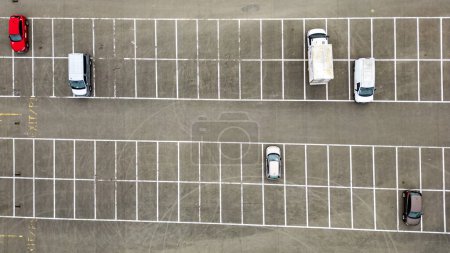 Aus der Luftperspektive erfasst das Bild die geordneten Muster eines Parkplatzes mit einer Handvoll Autos, die innerhalb der knackigen, weiß gesäumten Flächen auf grauem Asphalt parken. Die Palette der Fahrzeuge und