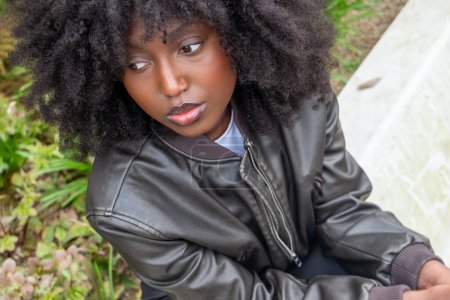 Eine nachdenkliche afroamerikanische Frau mit vollem Afro sitzt draußen, ihr distanzierter Blick und die lässige Pose vor der Gartenkulisse vermitteln einen Moment der Einsamkeit und inneren Besinnung. Umfassende Überlegungen