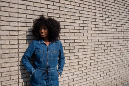 Una joven y elegante mujer afroamericana con un voluminoso peinado afro se encuentra casualmente contra una pared de ladrillo texturizado, con las manos metidas en los bolsillos de su elegante mono de mezclilla. Su mirada hacia los lados