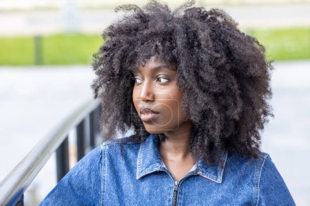 Esta imagen muestra a una joven afroamericana mirando a lo lejos. Ella está adornada con una elegante chaqueta de mezclilla azul con su voluminoso pelo negro texturizado en cascada alrededor de su cara, ofreciendo