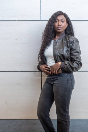 Una mujer africana equilibrada modela con confianza contra un muro de baldosas contemporáneo. Su chaqueta de cuero y jeans negros son el epítome de la elegancia urbana. Su cabello ondulado cae en cascada por sus hombros, completando el