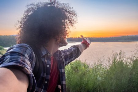 Eine Person mit lockigem Haar zeigt bei Sonnenuntergang auf einen See und verkörpert ein Gefühl von Frieden und Kontemplation. Lockige Person, die einen Sonnenuntergang am See genießt. Hochwertiges Foto