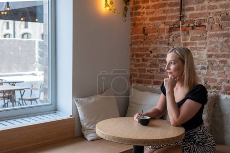 A contemplative young woman with blonde hair is seated in a rustic urban cafe, her gaze directed out a large window that frames the cityscape. She cradles a cup of coffee in her hands, lost in thought