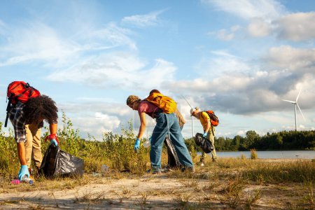 Four people engaged in a clean-up, picking up trash outdoors, wind turbine and lake in background, clear day. Volunteers Collecting Litter in Nature by Wind Turbine. High quality photo