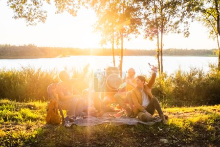 Eine Gruppe unterschiedlicher Erwachsener genießt ein Picknick in der Abenddämmerung am ruhigen Seeufer, das eine ungezwungene und fröhliche Atmosphäre widerspiegelt. Golden Sunset Picknick am See mit Freunden. Hochwertiges Foto