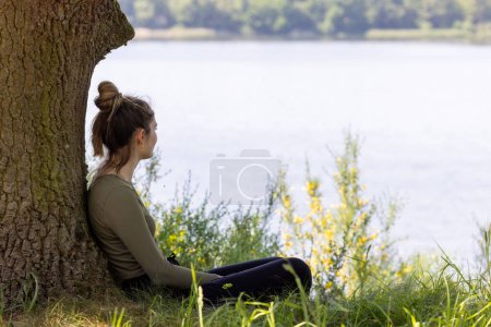 This photograph captures a young woman in a moment of peaceful solitude by the lake. She sits comfortably under the shade of an aged tree, overlooking the still waters. The scene is a blend of natural