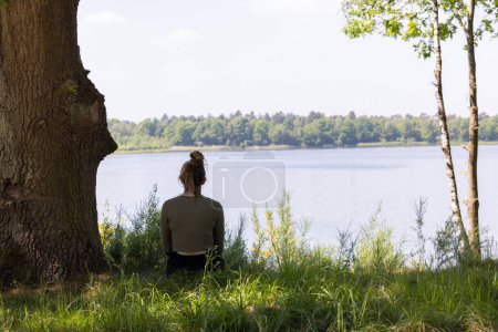 A woman is seen from behind, sitting thoughtfully by a serene lake, surrounded by the lush embrace of nature. The majestic tree to her left and the gentle waters before her create a naturally framed
