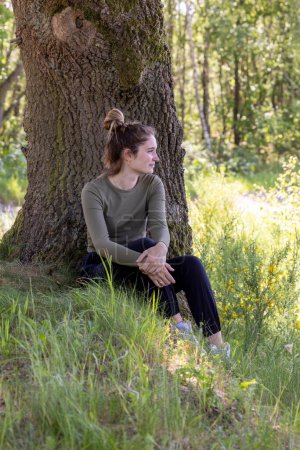 This image depicts a contemplative woman seated at the base of a large, textured tree in a verdant forest setting. Her gaze is directed off into the distance, suggesting introspection or a deep