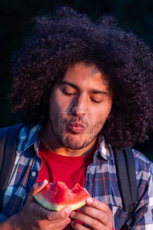 This image captures a young man with curly, dense afro hair enjoying a slice of juicy watermelon. He is of mixed ethnicity, likely Afro-Latino, with medium brown skin. His eyes are closed in delight
