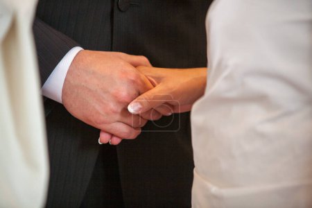 In an up-close and personal depiction of a wedding ceremony, the image focuses on the hands of partners as they exchange rings. The grooms black suit and the brides white dress provide a timeless