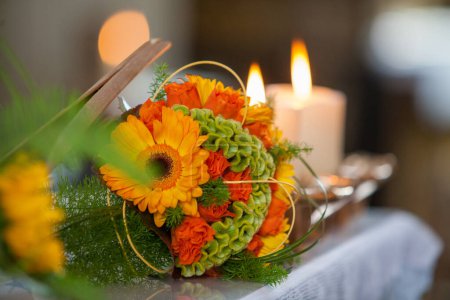 Una vibrante pieza central floral compuesta por flores de naranja y exuberante vegetación se asienta elegantemente sobre una mesa festivamente adornada, flanqueada por el suave resplandor de las velas encendidas. Esta configuración crea una cálida e invitadora