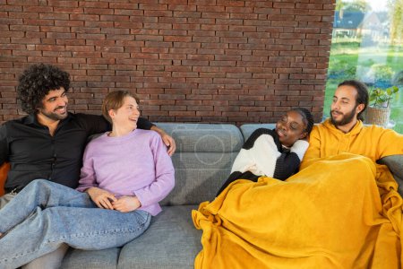 A diverse group of friends shares a warm, intimate moment on a gray couch, draped with a yellow blanket, in a living room with a brick wall, embodying comfort, friendship, and relaxation in a homey