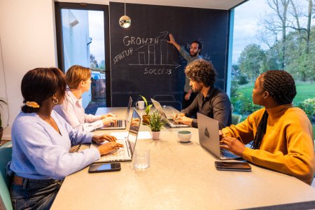 In a well-lit, modern office, a group of focused professionals sit around a table working on laptops, while a team member enthusiastically presents growth strategies on a chalkboard, embodying startup