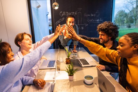 A diverse group of professionals share a high-five in a contemporary office setting, exuding enthusiasm and teamwork. Behind them, a chalkboard filled with business growth concepts speaks to their