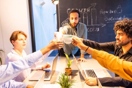 A team of young professionals in a well-lit office environment share a toast over coffee cups, marking a moment of success or agreement. The background chalkboard lists inspirational startup concepts