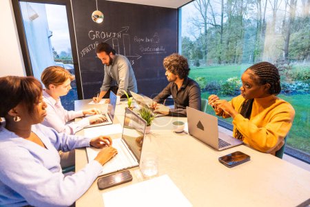 In a spacious modern workspace with a clear view of a lush garden, a focused and diverse team collaborates on a startups growth and sustainability plan. A blackboard in the background highlights key