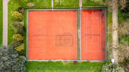 An aerial shot showcasing two adjacent red clay tennis courts, clearly marked for play and enclosed by vibrant green grass. The courts are devoid of players, highlighting their symmetry and the