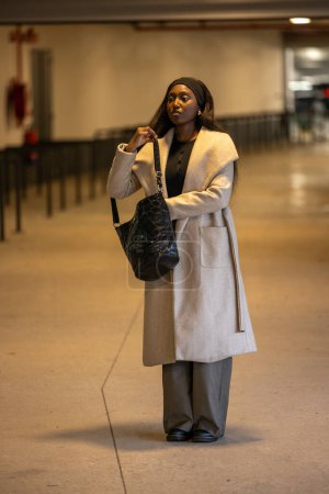 In einem geräumigen städtischen Korridor steht eine schwarze Afroamerikanerin in einem schicken beigen Mantel und hält eine schwarze Designerhandtasche in der Hand. Ihr besinnlicher Blick und die weiche Innenbeleuchtung betonen sie
