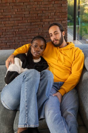 This image captures a poignant moment of a multiethnic couple enjoying a quiet embrace on a gray couch. Against a rich, brick wall background, the warm tones of the mans yellow hoodie contrast with