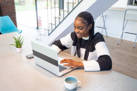 Capturing the essence of remote work, a young woman with braided hair smiles brightly as she types away on her laptop. The contemporary home setting, complete with stylish interior and natural light