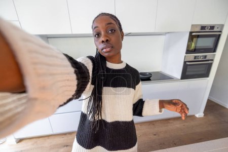 In diesem einladenden Bild streckt eine junge Frau ihren Arm aus, um ein Selfie in den engen Räumen einer modernen Küche zu machen. Ihr gestreifter Pullover verleiht dem minimalistischen Hintergrund einen Hauch von persönlichem Stil