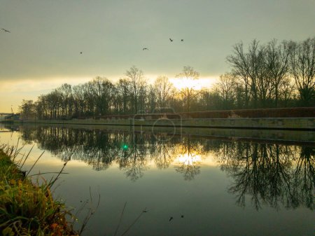Dieses Foto fängt die heitere Schönheit eines Flusses in der Morgendämmerung ein, wobei sich das goldene Licht der aufgehenden Sonne auf der Wasseroberfläche reflektiert. Vögel im Flug verleihen dem friedlichen Flussufer ein dynamisches Element