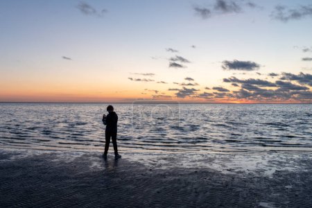 Eine einsame Person steht am Rande eines ruhigen Meeres und blickt in Richtung Horizont, wo ein sanfter Sonnenuntergang einen Farbteppich über den Himmel wirft, der sich subtil in der Wasseroberfläche widerspiegelt und eine