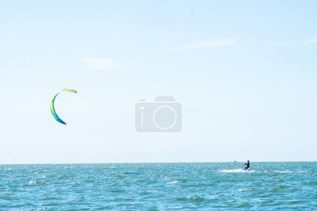 A kitesurfer harnesses the wind power, gliding across the seas surface under a vast, clear sky. The prominent kite contrasts against the blue backdrop, adding dynamism to the serene aquatic landscape