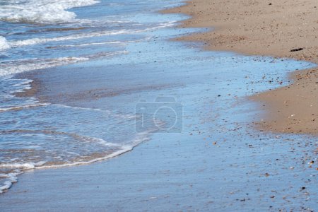 A gentle surf washes over a sandy beach, littered with shells and pebbles, under a clear blue sky. The intimate perspective highlights the tranquility of the seaside ambiance. Soft Waves Lapping at a