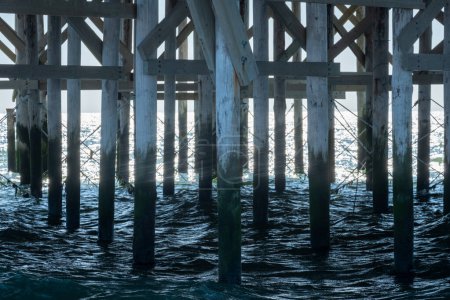Capturing the intricate patterns of light and shadow, this image showcases the robust wooden structure of a pier with its pillars and crossbeams plunging into the dark, swirling waters below, offering