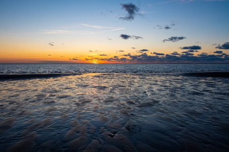 The sun sets over a rippled seashore, painting the clouds with warm hues and casting a peaceful glow on the textured sand, encapsulating the serene beauty of the coastal evening. Dramatic Sunset Over
