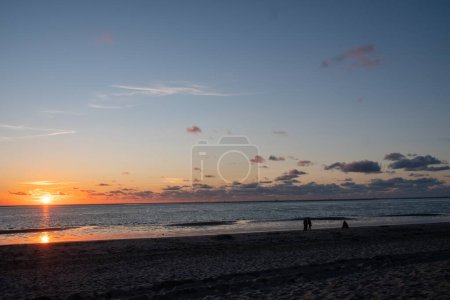 The image captures a picturesque sunset on the beach, with the sun dipping into the horizon. The expansive sky is strewn with soft clouds, and the calm sea reflects the fading sunlight. Two distant