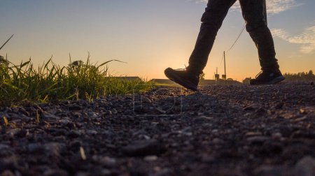 Cette image montre une vue en angle bas d'un individu jambes alors qu'ils marchent sur un chemin rural au coucher du soleil. La lumière dorée du soleil couchant crée des silhouettes frappantes et des ombres allongées