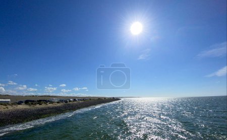 This vibrant image captures a bright sunny day at the seaside, featuring a glistening sea reflecting the sunlight, a clear blue sky, and a stony shoreline that leads to a distant horizon. The suns
