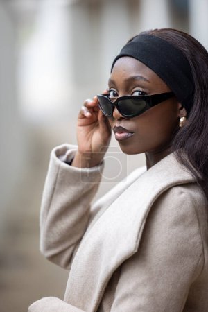 An elegant young woman peers over her sunglasses, exuding confidence and style. Her headband complements her chic beige coat, capturing a moment of urban sophistication. Sophisticated Young Woman in