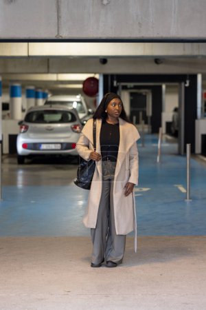A poised young woman walks confidently through a parking garage, dressed in a stylish beige coat over a black outfit, accessorizing with a headband and carrying a black handbag. Elegant Woman in Chic